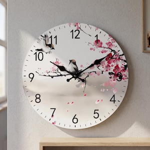 Puede incluir: Un reloj de pared redondo con esfera blanca y números arábigos negros. El diseño muestra dos pájaros pequeños posados en una rama oscura con flores de cerezo rosas. El reloj tiene manecillas negras para las horas, minutos y segundos.