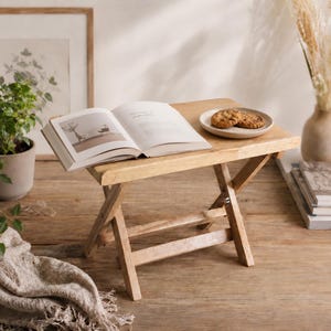 May include: A light-colored, wooden folding table with an open book and a small plate with two cookies. The table is on a wooden surface, with a plant and framed artwork in the background.