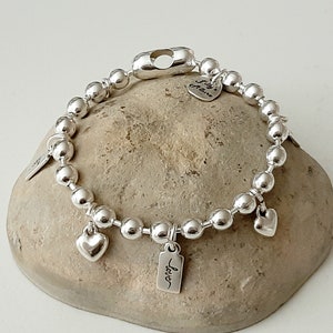 May include: Silver charm bracelet with a heart charm, a tag charm that says "Love", and a tag charm that says "Sally Grace". The bracelet is made of silver beads and has a lobster clasp.