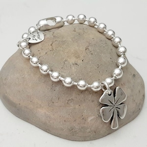 May include: Silver bracelet with a four-leaf clover charm. The bracelet is made of small silver beads and has a heart-shaped tag that says "Lilly's Jewelry".