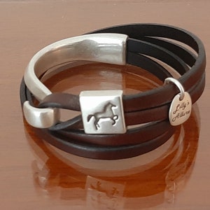 May include: A multi-strand leather bracelet with a silver-tone clasp and accents. The bracelet features a horse silhouette on the clasp and a small charm that reads "Lilly's Allure". The leather strands are black and brown.