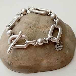 Könnte beinhalten: Silberarmband mit einem Toggle-Verschluss, ovalen Gliedern und kleinen runden Perlen. Das Armband hat einen herzförmigen Anhänger mit dem Text "Lilly's Allure".
