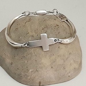May include: A silver bracelet with a cross pendant. The bracelet has a curved design with a clasp closure. The cross is centered on the bracelet and is a solid, rectangular shape. The bracelet is displayed on a stone.