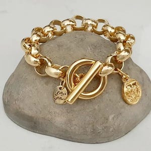 May include: A gold-colored bracelet with a toggle clasp and a chain-link design. The bracelet features a small charm with the text "Lilly's Atelier" and a round coin charm. The bracelet is displayed on a gray stone surface.