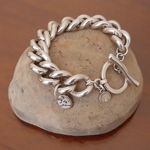 May include: Silver chain bracelet with a toggle clasp and a heart-shaped charm that says "Lilly's Allure".
