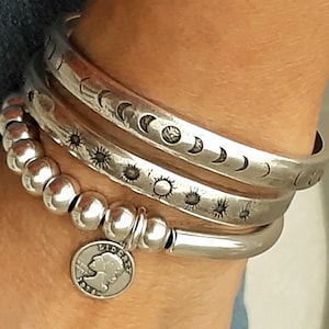 May include: Three silver bracelets with a moon and star design, stacked on a wrist. The bottom bracelet is a plain silver band. A silver charm with a coin design hangs from the bottom bracelet.