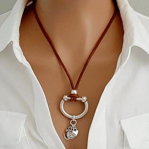 May include: A brown leather cord necklace with a silver-colored pendant. The pendant features a circular design with a heart and other charms. The necklace is worn over a white collared shirt.