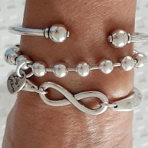 Könnte beinhalten: Eine Sammlung von Silberarmbändern. Das obere Armband ist ein Armreif mit Kugelenden. Darunter befindet sich ein Perlenarmband und unten ein Unendlichkeitszeichen-Armband. Die Armbänder werden zusammen getragen.