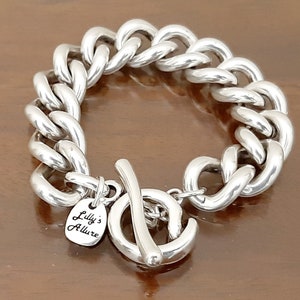 May include: Silver chain bracelet with a chunky curb link design. The bracelet has a toggle clasp and a small silver tag that reads "Lilly's Allure".