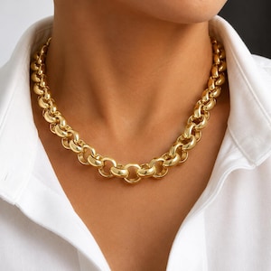 May include: A chunky gold-tone chain necklace featuring large, polished, circular interlocking links. The necklace rests around the neck, worn over a crisp white collared shirt. The metal surface is highly reflective, creating bright highlights.