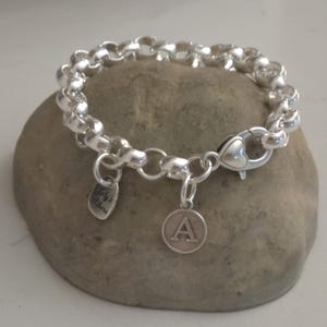 May include: A silver chain link bracelet featuring a heart-shaped lobster clasp, a small oval charm engraved with the text "Milly's Allure", and a circular charm displaying the capital letter "A" in a serif font, all crafted in a polished finish.