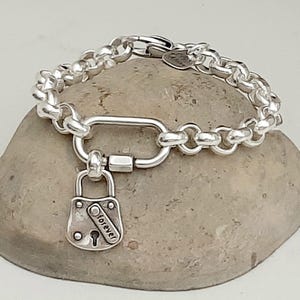 May include: A silver bracelet featuring a thick chain design and a large clasp. A small padlock charm with the word "forever" is attached. The bracelet is displayed on a grey stone.