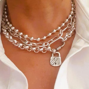 May include: A silver necklace featuring three chains, including a chain with small round beads, a chain with larger oval links, and a chain with a padlock pendant. The padlock has a keyhole and the word "forever" engraved on it.
