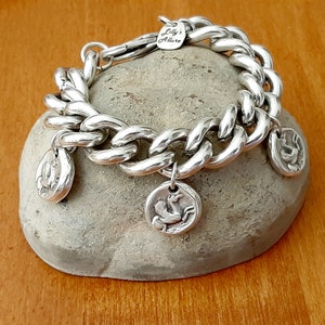 May include: A silver chain bracelet with three silver charms featuring a bird design. The bracelet is on a gray rock with the text "Lilly's Allure" on a tag attached to the bracelet.