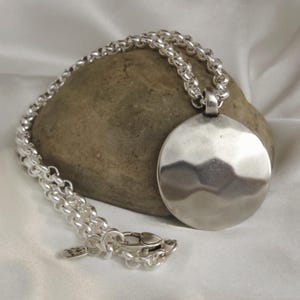 May include: A silver-toned necklace featuring a large, circular, hammered-finish pendant suspended from a thick, round link chain. The chain includes a lobster claw clasp and a small rectangular metal tag near the closure mechanism.