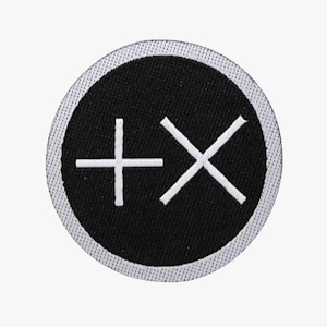 May include: A circular black patch with a white border. The patch features a white plus sign and an X symbol, both in a simple, bold style. The design is centered on the patch.