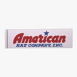 Könnte beinhalten: Ein rechteckiges weißes Etikett mit dem roten Wort "American", einem blauen Stern und dem blauen Text "HAT COMPANY, INC.". Das Etikett ist für eine Hut-Firma.