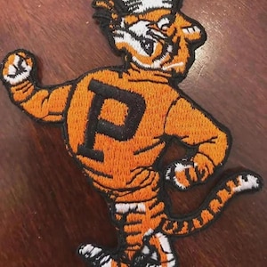 May include: Embroidered patch featuring a tiger mascot in orange, black, and white. The tiger is wearing a hat and a jersey with a large black "P". The patch is likely for a school or sports team.
