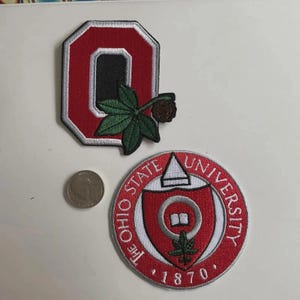 Puede incluir: Dos parches bordados: una "O" roja y negra con una hoja verde y una nuez marrón, y un parche circular con "THE OHIO STATE UNIVERSITY" y "1870" en rojo y blanco. Una moneda está al lado de los parches.