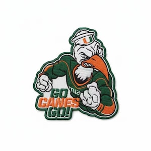 Puede incluir: Parche bordado con una mascota verde y naranja con una expresión decidida. La mascota lleva un sombrero y uniforme, con el texto "GO CANES GO!" en naranja y verde. El parche tiene un fondo blanco.
