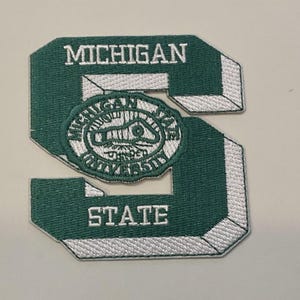 Könnte beinhalten: Ein grün-weißer, gestickter Aufnäher in Form des Buchstabens "S". Der Aufnäher zeigt die Wörter "MICHIGAN" und "STATE" in weißen Blockbuchstaben. Ein rundes Emblem mit den Worten "MICHIGAN STATE UNIVERSITY" befindet sich in der Mitte.