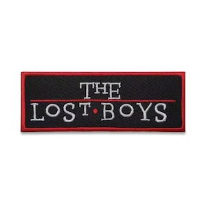 May include: A rectangular black patch with a red border. The text "THE LOST BOYS" is embroidered in white, with a red dot between the words. The patch is likely for clothing or accessories.