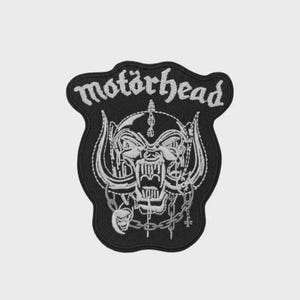 May include: Black embroidered patch featuring the word "motörhead" in white script above a detailed white illustration of a skull with tusks, horns, and chains. The patch has a black border.