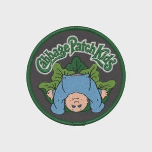 May include: Embroidered patch featuring a Cabbage Patch Kids design. The patch has a dark gray background with a green border. The design includes a baby in a blue outfit, surrounded by green leaves, and the words "Cabbage Patch Kids" in green.