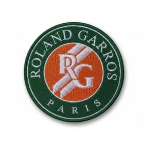 Peut inclure: Un écusson rond avec l'inscription "ROLAND GARROS PARIS" en blanc sur un fond vert. Le centre présente un cercle orange avec des rayures blanches et les lettres "RG".