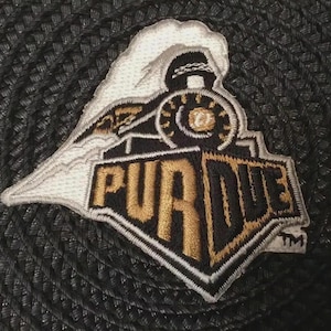 May include: Embroidered patch featuring a stylized train with the word "PURDUE" in gold and black. The patch has a white border and a steam cloud. The patch is on a black textured surface.
