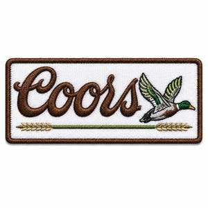 Puede incluir: Un parche rectangular bordado con fondo blanco y borde marrón. Presenta el texto marrón Coors en letra cursiva, un pato real volando con plumas verdes, blancas y marrones, y un gráfico horizontal de un tallo de trigo de color verde.