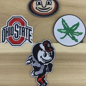 Puede incluir: Cuatro parches bordados con imágenes de la Universidad Estatal de Ohio. Incluye el logotipo de Ohio State, una hoja de Buckeye y dos versiones de la mascota de Ohio State, Brutus Buckeye. Los parches son rojos, blancos, negros y verdes.