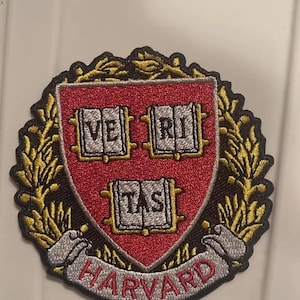 May include: Embroidered patch featuring the Harvard University crest. The crest is a shield shape with a red background and three open books, each with text. The word "HARVARD" is arched below the shield. The patch is trimmed with gold and black.