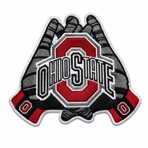Puede incluir: Un parche bordado que muestra dos guantes de fútbol americano grises y negros colocados juntos, con el logotipo rojo y blanco de Ohio State en el centro. El texto OHIO STATE está bordado en blanco sobre el centro de los guantes.