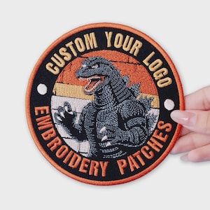 May include: Embroidered patch featuring a Godzilla design with the text "CUSTOM YOUR LOGO EMBROIDERY PATCHES". The patch has a black border with orange and white accents. The Godzilla illustration is in shades of gray.