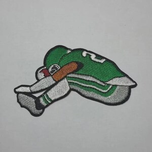 May include: Embroidered patch of a football player in a green uniform with white and grey details. The patch shows the player kneeling, head bowed. The patch is on a white background.