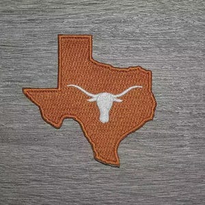Peut inclure: Écusson brodé en forme d'État du Texas, de couleur orange, avec une silhouette de longhorn blanche au centre. Sur un fond gris à effet de grain de bois.