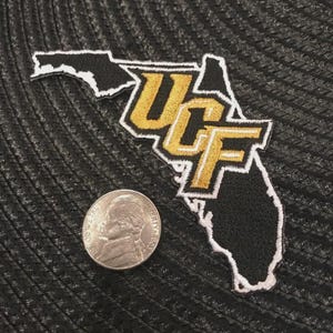 May include: A black and gold embroidered patch in the shape of Florida with "UCF" in gold lettering. A nickel is next to the patch for size comparison. The patch has a white border and is on a textured black surface.