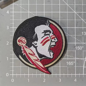 May include: Embroidered patch featuring a Seminole head in profile, with white, red, and black colors. The patch is circular, with a black background and a red border. The Seminole's face is white with red accents.