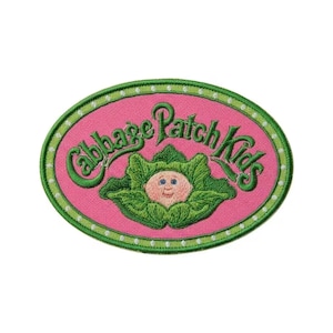May include: An oval-shaped embroidered patch featuring the text "Cabbage Patch Kids" in green script. The patch has a pink background with a green border and a cartoon baby face in a cabbage.