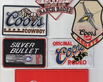 May include: Collection of embroidered patches featuring the Coors logo and related designs. Patches include the words "Banquet", "Cowboy", "Silver Bullet", and "Rodeo". Colors include red, white, blue, and black.