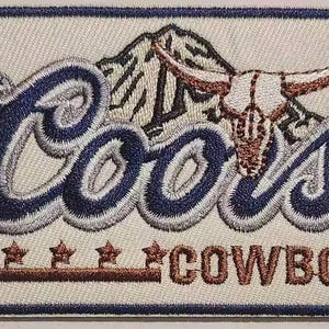 Puede incluir: Parche bordado con la palabra "Coors" en azul y blanco, con un diseño de montaña y calavera de toro. La palabra "COWBOY" está bordada en marrón debajo, con acentos de estrella. El parche tiene un borde azul.
