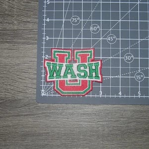 May include: Embroidered patch with the word "WASH" in green letters on a red and white background. The patch is in a shield shape and is on a gray cutting mat.