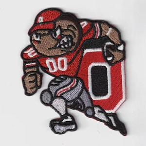 May include: Embroidered patch depicting a cartoon football player in a red uniform with the number "00". The player is running with a large red "O" on the side. The patch has a white background.