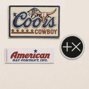 May include: Three embroidered patches. The top patch reads "Coors Cowboy" with a bull skull graphic. The middle patch says "American Hat Company, Inc." The bottom patch is a black circle with a white plus and X symbol.