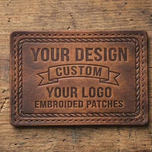 May include: A rectangular, brown leather patch with the words "YOUR DESIGN CUSTOM YOUR LOGO EMBROIDERED PATCHES" embossed on it. The patch has a stitched border and is set against a wooden background.