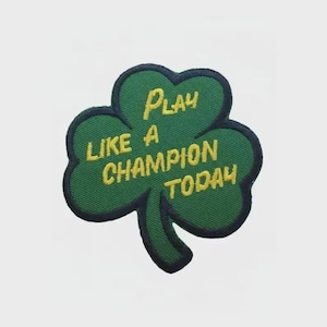 Könnte beinhalten: Grüner Kleeblatt-Aufnäher mit dem Text "Play Like a Champion Today" in Gelb. Der Aufnäher hat einen dunkelgrünen Rand. Geeignet für Kleidung oder Accessoires.