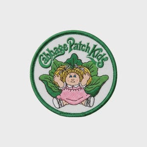 May include: Embroidered patch with the Cabbage Patch Kids logo. It shows a doll in a pink dress, surrounded by green leaves, with the words "Cabbage Patch Kids" in green script. The patch has a white background and a green border.