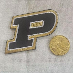 May include: A black and gold embroidered patch in the shape of the letter "P" with a gold coin featuring an angel. The patch has a white border and gold stitching. The coin is gold-colored and has an image of an angel.