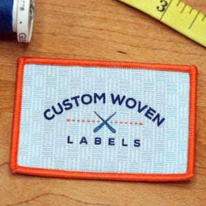 Może przedstawiać: Prostokątna tkana etykieta z pomarańczową obwódką i jasnoniebieskim tłem. Na etykiecie widnieje napis "CUSTOM WOVEN LABELS" w kolorze granatowym, z grafiką skrzyżowanych nożyczek. Szpulka nici i miarka znajdują się w tle.